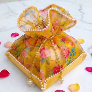 Könnte beinhalten: Ein quadratischer gelber Geschenkbeutel aus Stoff mit einem Blumenmuster aus rosa und roten Rosen. Der Beutel besteht aus transparentem Organzastoff, ist an den Rändern mit weißen Kunstperlen verziert und hat goldene Kordelzüge mit Quasten.