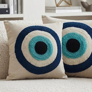 May include: Two square throw pillows with a beige fabric base, each featuring a centered, circular evil eye motif. The design consists of concentric rings in dark blue, bright turquoise, and a solid black center, created with a textured tufted finish.