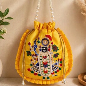 Puede incluir: Un bolso amarillo con cordón y un diseño de deidad bordado. El bolso presenta un asa de cuentas y un borde con volantes. La deidad está representada con detalles coloridos, incluyendo acentos rojos, azules y verdes. El bolso cuelga de un gancho decorativo.