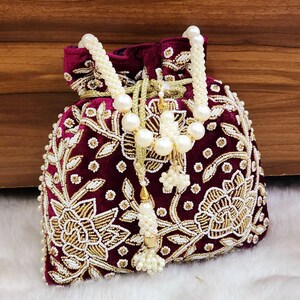 Può includere: Un astuccio in velluto bordeaux con ricami floreali dorati e bianchi. La chiusura con coulisse è ornata da un nappa di perle e oro. La borsa presenta un manico di perline ed è progettata per occasioni speciali.