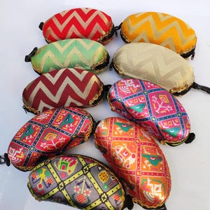 Puede incluir: Una colección de estuches para gafas estampados y coloridos. Los estuches presentan varios diseños, incluyendo zigzags y patrones geométricos, en tonos de rojo, amarillo, verde y rosa. Cada estuche tiene una cremallera negra y ribete dorado, de unos 16 cm de largo.