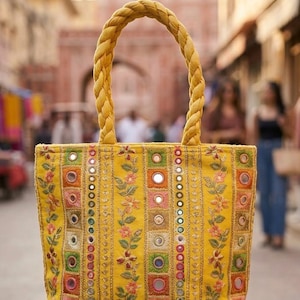 Könnte beinhalten: Eine gelbe Tragetasche mit geflochtenem Griff. Die Tasche ist mit bunten, gestickten Blumenmustern, kleinen Spiegeln und quadratischen und kreisförmigen Akzenten verziert. Der Hintergrund zeigt eine verschwommene Straßenszene.