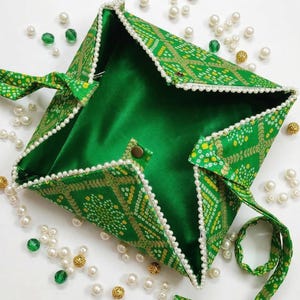 Può includere: Una borsa quadrata in tessuto verde con un motivo dorato e giallo. La borsa è rifinita con perle bianche e ha un interno in raso verde. Un laccio verde abbinato chiude la borsa e si vede un bottone dorato.