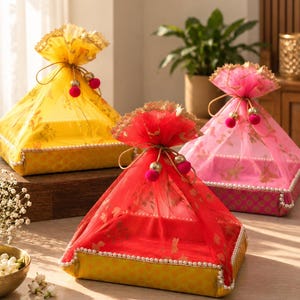 May include: Three square gift trays featuring fabric covers in yellow, red, and pink. Each tray has a base with a patterned texture, a sheer mesh top with gold floral accents, a pearl-beaded border, and a drawstring closure with pom-pom details.