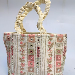 Könnte beinhalten: Cremefarbene Tragetasche mit geflochtenem Griff. Die Tasche hat vertikale Streifen mit floralen Stickereien und quadratischen Spiegelakzenten in Rosa, Grün und Gold. Die Griffe sind aus einem gedrehten, cremefarbenen Stoff.