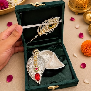 Puede incluir: Una caja de terciopelo verde oscuro que contiene un broche plateado en forma de pluma con cristales transparentes y un acento de ojo de pavo real, junto a un recipiente plateado en forma de hoja con tallo dorado, cristales y una piedra roja.