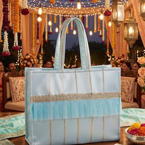 Può includere: Una borsa tote azzurra con strisce verticali dorate, un bordo orizzontale ricamato in oro e una balza in tulle azzurro plissettato al centro. La borsa presenta due manici superiori coordinati e una forma rettangolare strutturata.