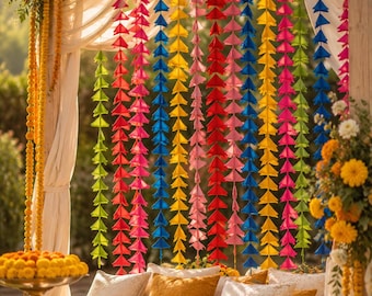 May include: A wooden frame structure draped with white fabric and adorned with vertical garlands of colorful fabric triangles in red, pink, blue, yellow, and green. A gold pedestal bowl holds yellow marigold flowers near white and gold pillows.
