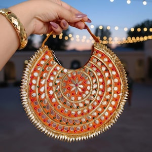 May include: An orange, half-moon clutch bag with detailed embroidery. The bag features a central floral design, surrounded by concentric circles of silver, red, and green embellishments. The edges are trimmed with gold and silver details.