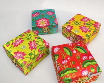 May include: Four square decorative boxes with floral patterns. The boxes are in various colors, including teal, yellow, and red, and feature pink lotus flowers and green leaves. Each box has a gold-colored clasp.