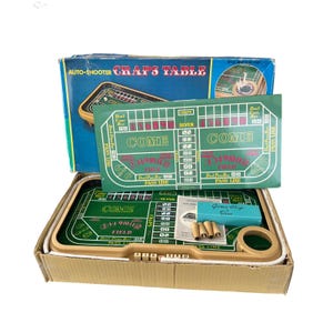 May include: A vintage craps table game set. The box is open, revealing a green felt table with white markings and the words "COME" and "PASS LINE." The set includes dice, chips, and a blue dice container. The box reads "AUTO-SHOOTER CRAPS TABLE."