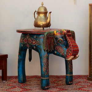 May include: A hand-painted blue elephant-shaped side table with intricate floral designs in red, gold, and green. A gold teapot sits on the table's round top. The table is on a red patterned rug.