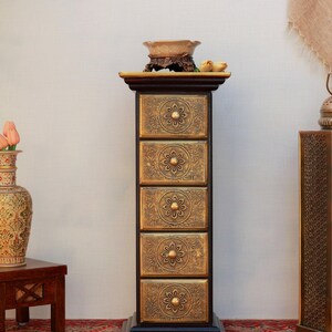 May include: A tall, dark wood cabinet with five drawers, each adorned with a gold floral design and a round knob. The cabinet is topped with a decorative bowl and sits on a patterned rug, showcasing an antique aesthetic.