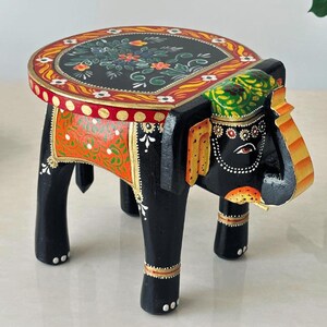 May include: A hand-painted wooden elephant stool. The black elephant-shaped base supports a round tabletop decorated with floral patterns in red, green, and yellow. The stool is approximately 12 inches tall.