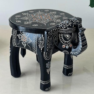 May include: A black wooden elephant stool with a round top. The top is decorated with a central floral design in orange, white, and blue. The elephant's body and legs are intricately painted with white floral patterns. The stool is approximately 12 inches tall.