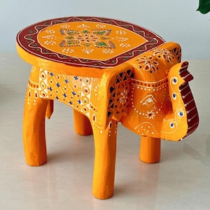 May include: A hand-painted wooden elephant stool in shades of orange, red, and white. The stool features intricate floral patterns on the seat and body. The elephant-shaped legs provide support. A decorative piece for any room.