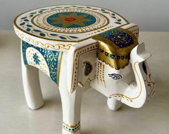 May include: A white, hand-painted wooden elephant stool with intricate gold and blue floral designs. The round top features a central floral motif. The elephant's head is adorned with gold accents, and the legs are painted with gold dots.