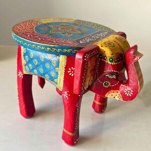 May include: A hand-painted wooden elephant stool. The stool features a red body, blue and yellow accents, and intricate floral patterns. The elephant's head forms the front of the stool, with the trunk extending downwards.