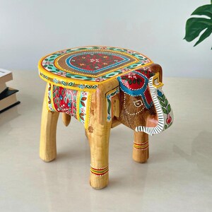 May include: A hand-painted wooden elephant stool with a colorful floral design on the seat. The elephant's head and body are brown, with painted details in red, blue, and yellow. The legs are light wood, and the stool is approximately 12 inches tall.
