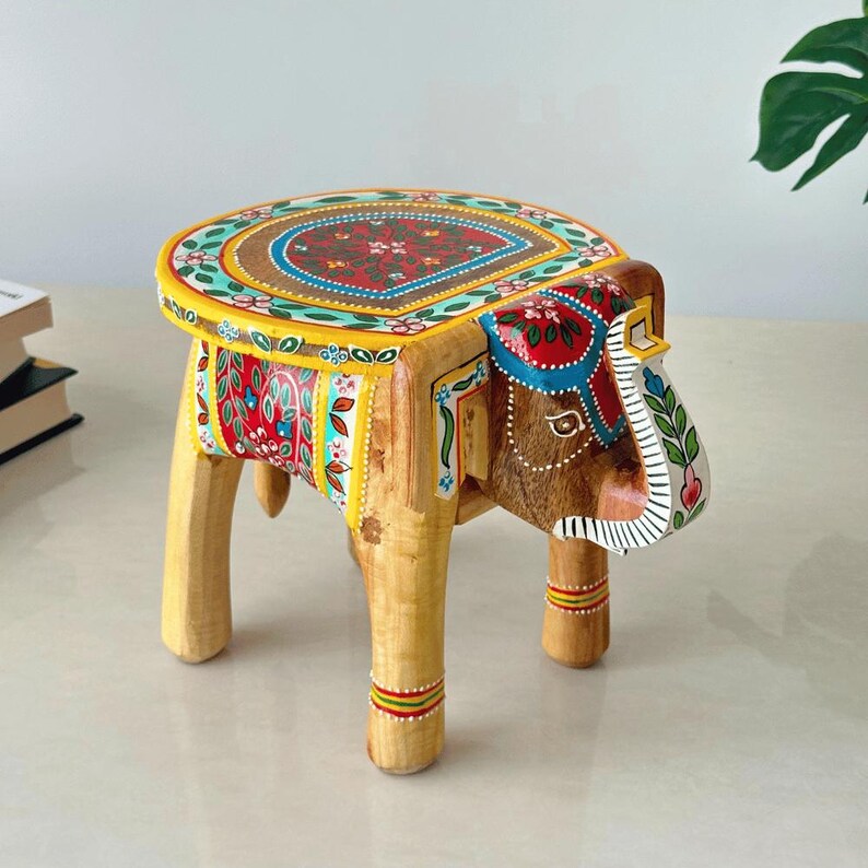 May include: A hand-painted wooden elephant stool with a colorful floral design on the seat. The elephant's head and body are brown, with painted details in red, blue, and yellow. The legs are light wood, and the stool is approximately 12 inches tall.