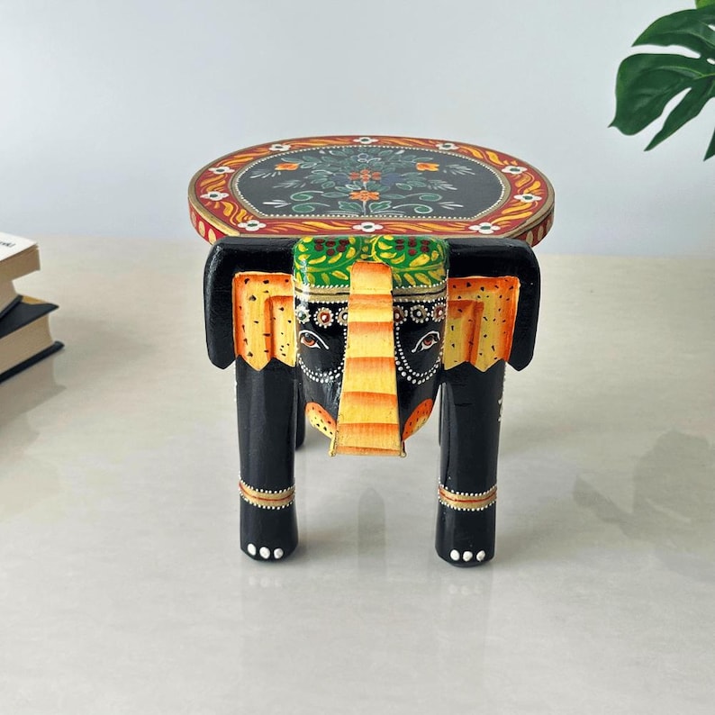 May include: A decorative wooden elephant stool. The black stool features an oval top with floral designs and an elephant head design. The elephant's head is painted with yellow, green, and orange accents. The legs are black with gold details.