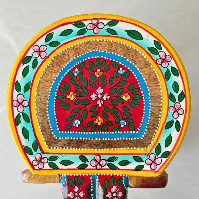 May include: A hand-painted wooden decorative piece with a semi-circular top. The design features a red center with floral patterns, surrounded by blue, yellow, and green borders. Pink flowers and green leaves adorn the edges. The piece is supported by a wooden base.