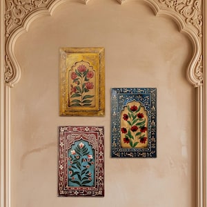 May include: Three framed floral paintings in yellow, blue, and maroon hang on a beige wall. Each painting features a central floral design within an arched frame. The wall is adorned with an ornate, carved archway.