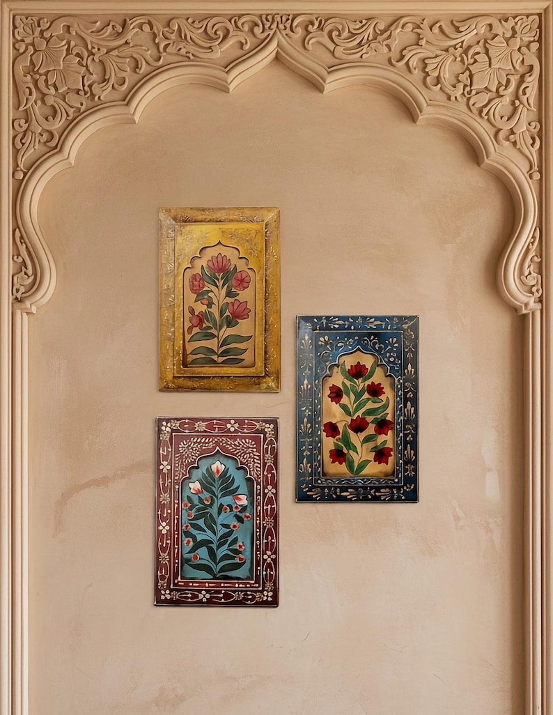 May include: Three framed floral paintings in yellow, blue, and maroon hang on a beige wall. Each painting features a central floral design within an arched frame. The wall is adorned with an ornate, carved archway.