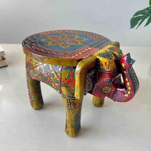 May include: A hand-painted wooden elephant stool. The stool features a round top with a colorful floral design in blue, red, and yellow. The elephant's body is painted in yellow and red with intricate floral patterns. The stool is approximately 12 inches tall.