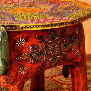 May include: A hand-painted, round wooden side table with intricate, colorful patterns. The tabletop features a mosaic of orange, yellow, purple, and blue designs. The red base is adorned with floral and geometric motifs, creating a vibrant, artistic piece.