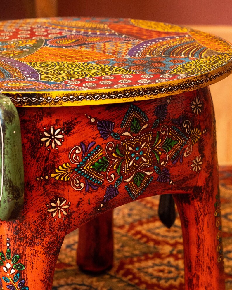 May include: A hand-painted, round wooden side table with intricate, colorful patterns. The tabletop features a mosaic of orange, yellow, purple, and blue designs. The red base is adorned with floral and geometric motifs, creating a vibrant, artistic piece.