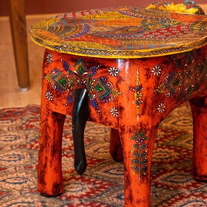 May include: A hand-painted orange wooden elephant stool with a round top. The top features a colorful paisley design. The stool has four legs and a black tail. The stool is approximately 15 inches tall.