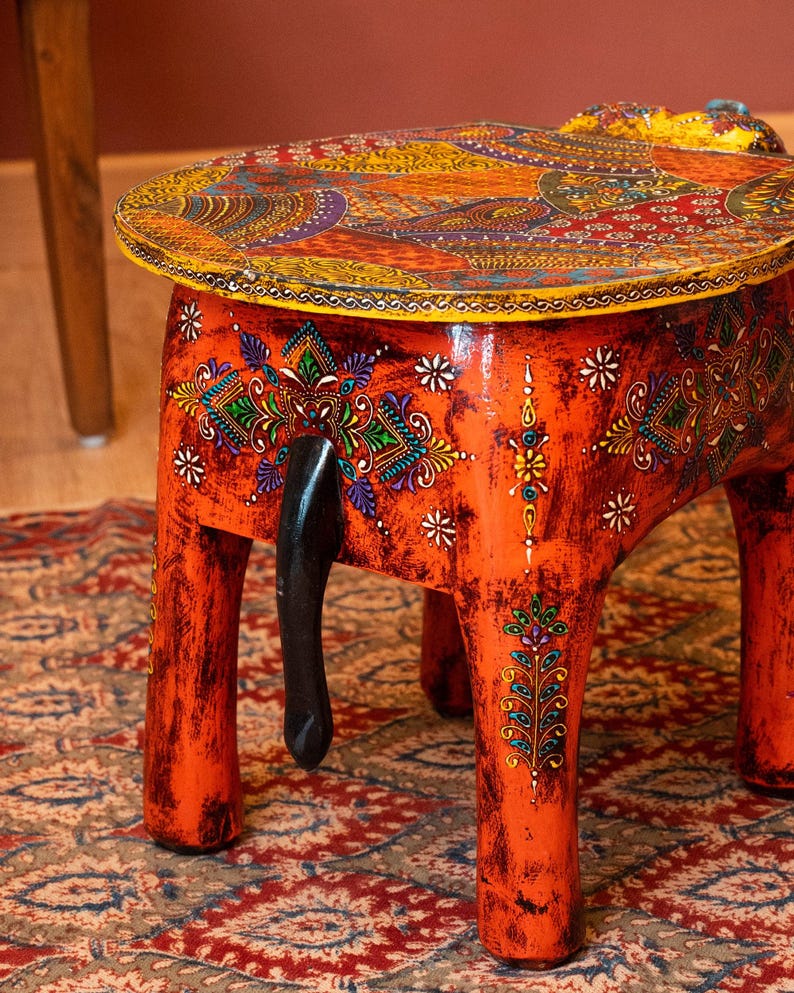 May include: A hand-painted orange wooden elephant stool with a round top. The top features a colorful paisley design. The stool has four legs and a black tail. The stool is approximately 15 inches tall.