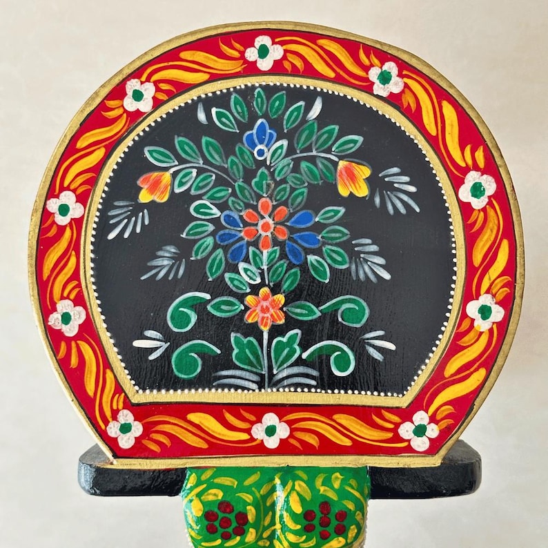 May include: A hand-painted, decorative wooden piece featuring a black, semi-circular design with a floral motif. The flowers are in shades of orange, blue, and red, with green leaves. The border is red and yellow with white flower accents.