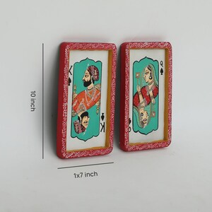 K&ouml;nnte beinhalten: Zwei rechteckige Wandkunstwerke, jeweils 25 cm hoch und 2,5x18 cm breit, die Spielkartenmotive darstellen. Die roten Rahmen haben ein wei&szlig;es Blumenmuster. Eines zeigt einen K&ouml;nig in Orange und das andere eine K&ouml;nigin in Rot, beide vor einem t&uuml;rkisfarbenen Hintergrund.