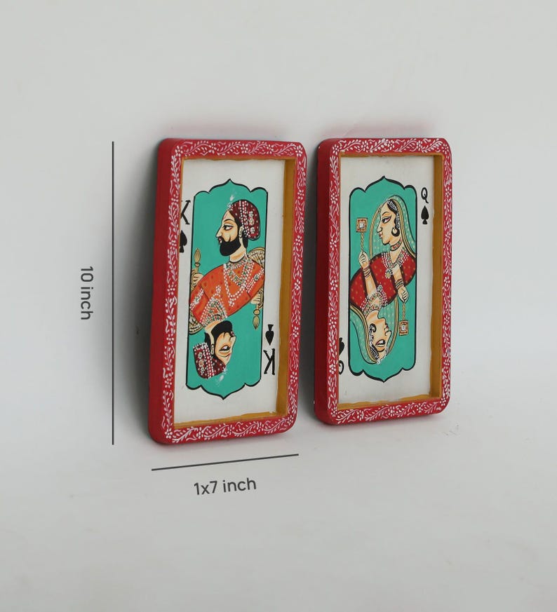K&ouml;nnte beinhalten: Zwei rechteckige Wandkunstwerke, jeweils 25 cm hoch und 2,5x18 cm breit, die Spielkartenmotive darstellen. Die roten Rahmen haben ein wei&szlig;es Blumenmuster. Eines zeigt einen K&ouml;nig in Orange und das andere eine K&ouml;nigin in Rot, beide vor einem t&uuml;rkisfarbenen Hintergrund.