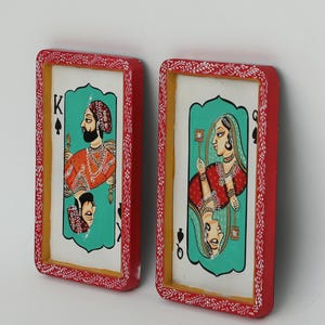 K&ouml;nnte beinhalten: Zwei gerahmte Spielkarten-Kunstwerke. Eines zeigt einen K&ouml;nig in Orange und Gold, das andere eine K&ouml;nigin in Rot und Gold. Beide Karten haben einen t&uuml;rkisfarbenen Hintergrund und sind rot mit wei&szlig;en floralen Details gerahmt.
