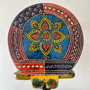 May include: A round, hand-painted wooden decorative piece with a vibrant floral design. The central flower is yellow and red against a blue background. The border features red, blue, and white patterns. The base is yellow with floral accents.