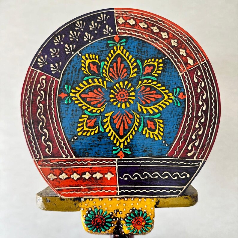 May include: A round, hand-painted wooden decorative piece with a vibrant floral design. The central flower is yellow and red against a blue background. The border features red, blue, and white patterns. The base is yellow with floral accents.