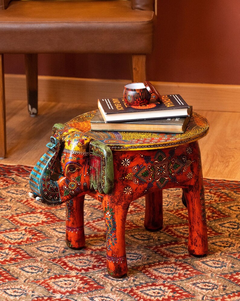 May include: A hand-painted, orange elephant-shaped side table with intricate floral and paisley designs. The table has a round top and four legs. Books and a small cup sit on top of the table. The table is on a patterned rug.