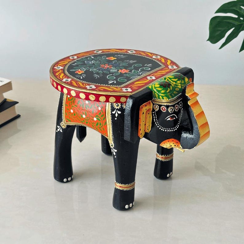 May include: A hand-painted wooden elephant stool. The black elephant-shaped base supports a round tabletop decorated with floral patterns in red, green, and yellow. The stool is approximately 12 inches tall.