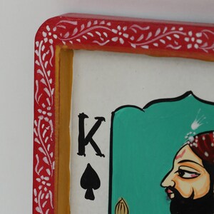 K&ouml;nnte beinhalten: Nahaufnahme einer Spielkarte mit dem K&ouml;nig der Pik. Die Karte hat einen roten Rand mit wei&szlig;en floralen Designs, einen gelben Innenrand und einen wei&szlig;en Hintergrund. Der K&ouml;nig ist im Profil mit Bart und Turban dargestellt.