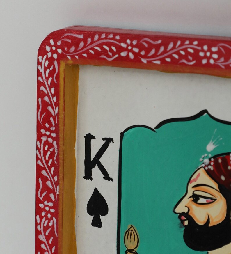 K&ouml;nnte beinhalten: Nahaufnahme einer Spielkarte mit dem K&ouml;nig der Pik. Die Karte hat einen roten Rand mit wei&szlig;en floralen Designs, einen gelben Innenrand und einen wei&szlig;en Hintergrund. Der K&ouml;nig ist im Profil mit Bart und Turban dargestellt.