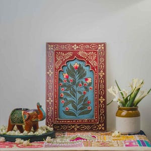 May include: A hand-painted wooden panel with a floral design in a red frame. The panel has a blue background with a painted tree and red flowers. An elephant candle holder and a vase of white flowers are also present.