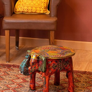 May include: A handcrafted, red elephant-shaped stool with intricate, colorful patterns. The stool features a round top and four legs, all adorned with detailed designs. The elephant's head and ears are painted with vibrant hues, adding to its decorative appeal. The stool is placed on a patterned rug.