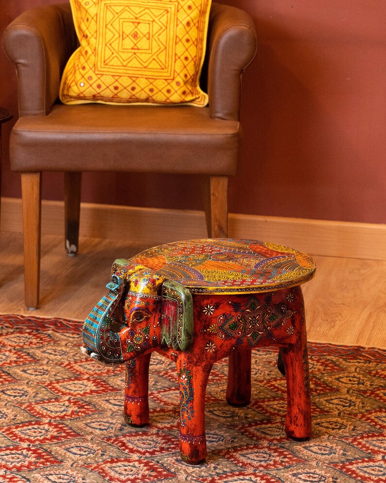 May include: A handcrafted, red elephant-shaped stool with intricate, colorful patterns. The stool features a round top and four legs, all adorned with detailed designs. The elephant's head and ears are painted with vibrant hues, adding to its decorative appeal. The stool is placed on a patterned rug.