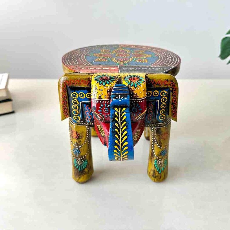May include: A hand-painted wooden elephant-shaped stool. The stool features a round top with a colorful floral design. The elephant's body is painted in yellow, red, and blue with intricate patterns. The legs are also painted yellow.