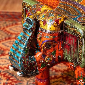 May include: A vibrantly painted wooden elephant sculpture, featuring intricate floral and paisley patterns in orange, yellow, blue, and green. The elephant's trunk and ears are adorned with detailed designs, showcasing a blend of colors and textures.