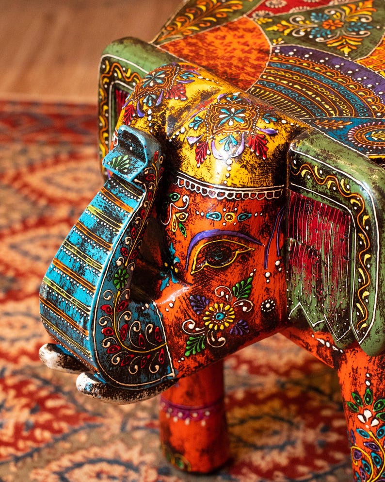 May include: A vibrantly painted wooden elephant sculpture, featuring intricate floral and paisley patterns in orange, yellow, blue, and green. The elephant's trunk and ears are adorned with detailed designs, showcasing a blend of colors and textures.