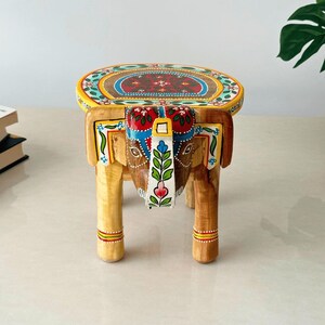 May include: A hand-painted wooden elephant stool with a round top. The top features a colorful floral design with red, blue, and yellow accents. The elephant's head and legs are intricately carved and painted with similar patterns.